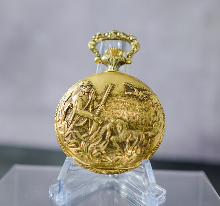 A gold pocket watch on a clear stand Description automatically generated